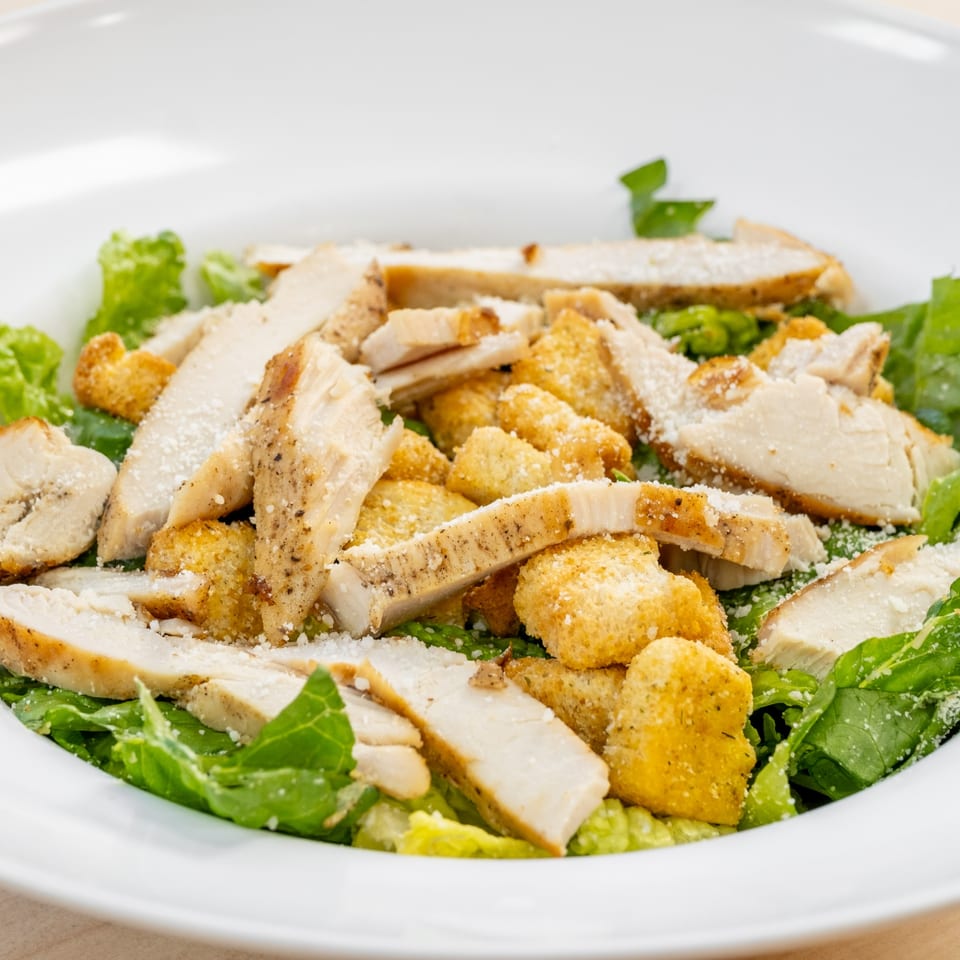 Close-up of a Caesar salad with grilled chicken, croutons, and grated cheese in a white bowl.