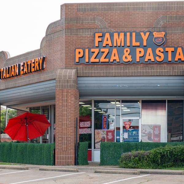 Exterior of Family Pizza & Pasta Italian Eatery, a brick building with a red umbrella and green hedge.
