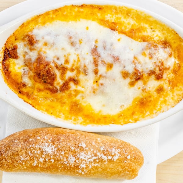 Baked ziti or lasagna in a white dish with melted cheese, served with a breadstick.