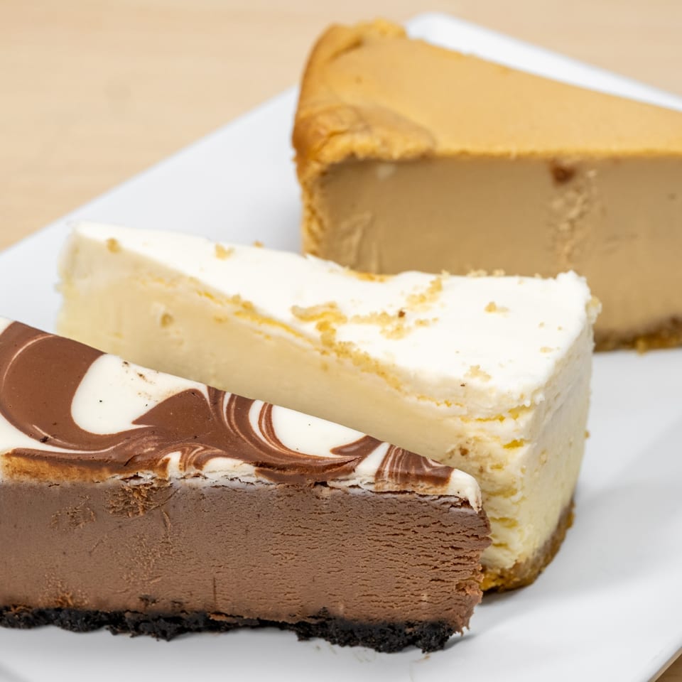 Three slices of cheesecake: chocolate swirl, classic vanilla, and caramel, on a white plate.